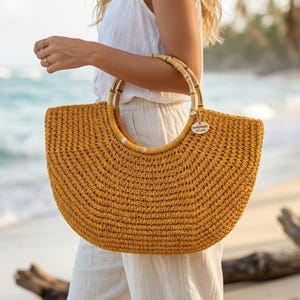 May include: Mustard yellow woven tote bag with bamboo handles. The bag has a rounded shape and a tag that reads "HANDMADE WITH LOVE". The bag is being held on a beach with the ocean in the background.