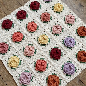 May include: A cream-coloured, crocheted blanket with a grid pattern and colourful, 3D crocheted flowers. The flowers are in shades of red, orange, yellow, pink, and purple, each surrounded by green leaves. The blanket is square.