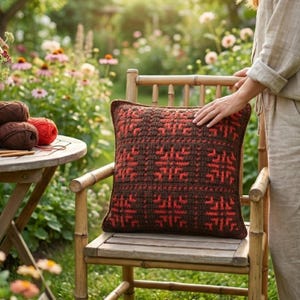 May include: A square, brown and red crocheted pillow with a geometric pattern rests on a bamboo chair. The pillow is in a garden setting, with yarn and knitting needles on a small table nearby. The pillow's dimensions are approximately 16 inches by 16 inches.