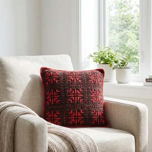 May include: A decorative square pillow with a dark brown and red geometric pattern. The pillow has a red border and is placed on a beige armchair with a knitted throw. The scene is lit by natural light from a nearby window.