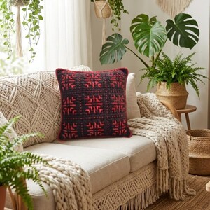 May include: A decorative square pillow with a red and black geometric pattern sits on a beige sofa. The pillow has a red border. A cream-colored knitted throw blanket and macrame accents add to the cozy, bohemian aesthetic.