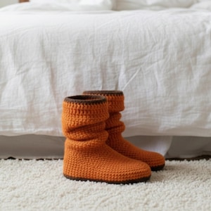 Puede incluir: Un par de botas de ganchillo naranjas con ribete marrón. Las botas están hechas de un hilo texturizado y tienen un diseño holgado. Se exhiben sobre una alfombra blanca frente a una cama blanca.