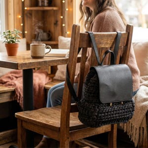 Peut inclure: Sac à dos en crochet noir avec rabat en cuir et bretelles réglables. Le sac est accroché à une chaise en bois. L'arrière-plan comprend une table en bois, une tasse de café et une plante. Le rabat a des ferrures dorées.