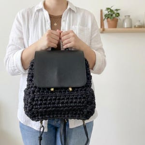 May include: Black crochet backpack with a flap closure. The top flap is made of smooth black material, and the bag has gold-coloured hardware. The backpack has adjustable straps and a handle.