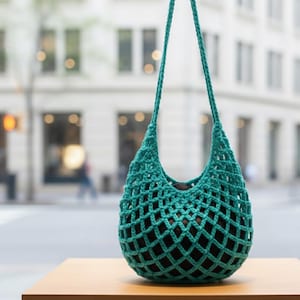 Könnte beinhalten: Eine türkisgrüne gehäkelte Handtasche mit netzartigem Design. Die Tasche hat eine runde Form und lange Träger zum Über-die-Schulter-Tragen. Das Innere der Tasche scheint eine dunkle Farbe zu haben, die mit dem türkisfarbenen Äußeren kontrastiert. Die Tasche steht auf einer Holzoberfläche.