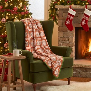 May include: A patterned throw blanket in shades of orange and white, draped over a green armchair. The blanket has a snowflake design. A white mug sits on a small wooden table. Christmas decorations are in the background.