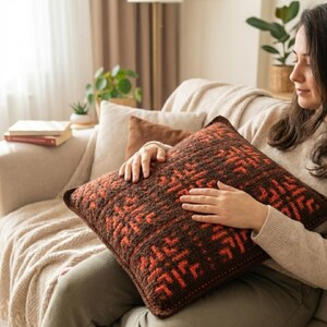 May include: A decorative square pillow with a geometric pattern in shades of brown and orange. The pillow is made of a textured fabric and appears to be handmade. The pillow is being held by a person sitting on a beige sofa.