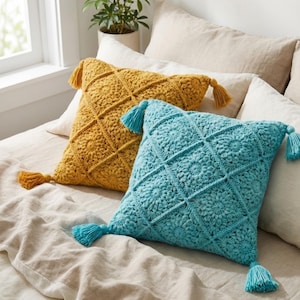 May include: Two square crochet pillows with diamond patterns and tassels. One pillow is mustard yellow, the other is turquoise. Both pillows are on a bed with neutral-colored bedding. The pillows add a decorative touch to the bedroom.