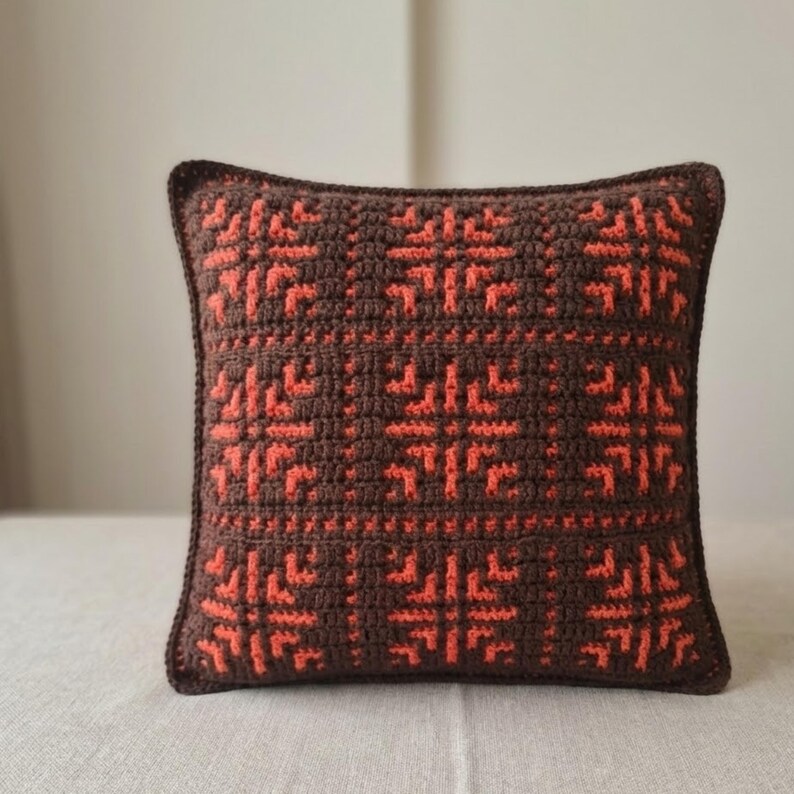 May include: A square crochet pillow with a brown background and a repeating orange geometric pattern. The pillow has a dark brown border and is likely used for home decor.