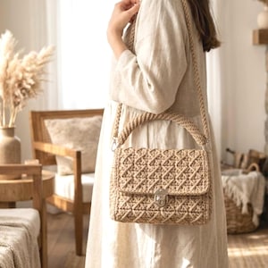 May include: A beige, hand-crocheted shoulder bag with a geometric pattern. The bag features a flap closure with a silver-colored clasp and a braided handle and strap. The bag is made of a soft, natural-looking yarn.