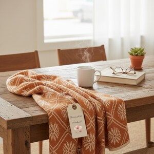 May include: An orange and cream throw blanket with a floral design is draped over a wooden table. A white mug with steam rising, a book, glasses, and a small potted plant are also on the table. A tag reads "Becca Arts Handmade."