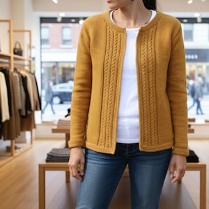 Puede incluir: Cárdigan amarillo mostaza con diseño de punto trenzado en los paneles delanteros. El suéter abierto se combina con una camiseta blanca y vaqueros azules. El cárdigan tiene mangas largas y puños acanalados. El fondo muestra una tienda de ropa.