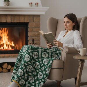 May include: A person sits in a beige armchair, reading a book, covered by a green and white patterned blanket. A lit fireplace is in the background, and a small wooden table holds a white mug.