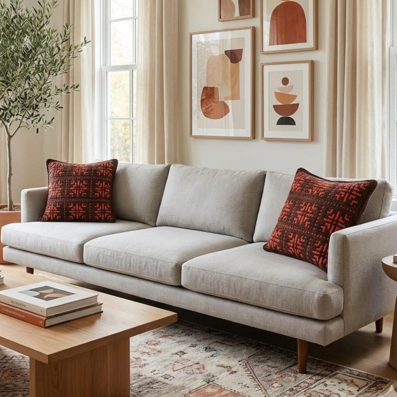 May include: A light gray three-cushion sofa with two decorative throw pillows. The pillows are a dark red with a geometric pattern. A wooden coffee table with books sits in front of the sofa. Artwork hangs on the wall.