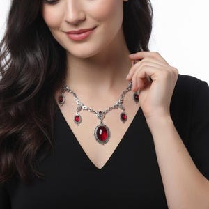 Peut inclure: Un collier argenté orné de pierres rouges de formes ovales et rondes. Le collier présente un design orné, avec un grand pendentif central et de plus petites pierres. La femme porte une robe noire.