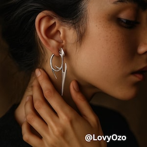 以下が含まれることがあります： 銀色のイヤリングを身につけた人のクローズアップ。イヤリングは、曲線と尖った要素を持つユニークな抽象的なデザインです。イヤリングは明るい銀色で、金属製のように見えます。画像には「@LovyOzo」というテキストが含まれています。