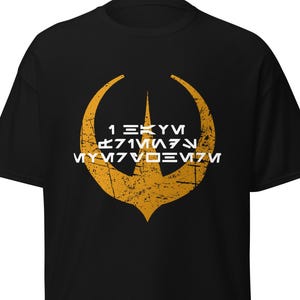 Może przedstawiać: Czarna koszulka z wytartą złotą grafiką i białym tekstem. Grafika to stylizowany emblemat z centralnym punktem i zakrzywionymi ramionami. Tekst jest w obcym języku. Koszulka wykonana jest z miękkiego materiału.