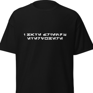 Può includere: T-shirt nera con testo bianco in un font stilizzato. Il testo sembra essere una frase o una citazione in una lingua unica, forse fittizia. La maglietta è un modello girocollo di base, adatta per l'abbigliamento casual.