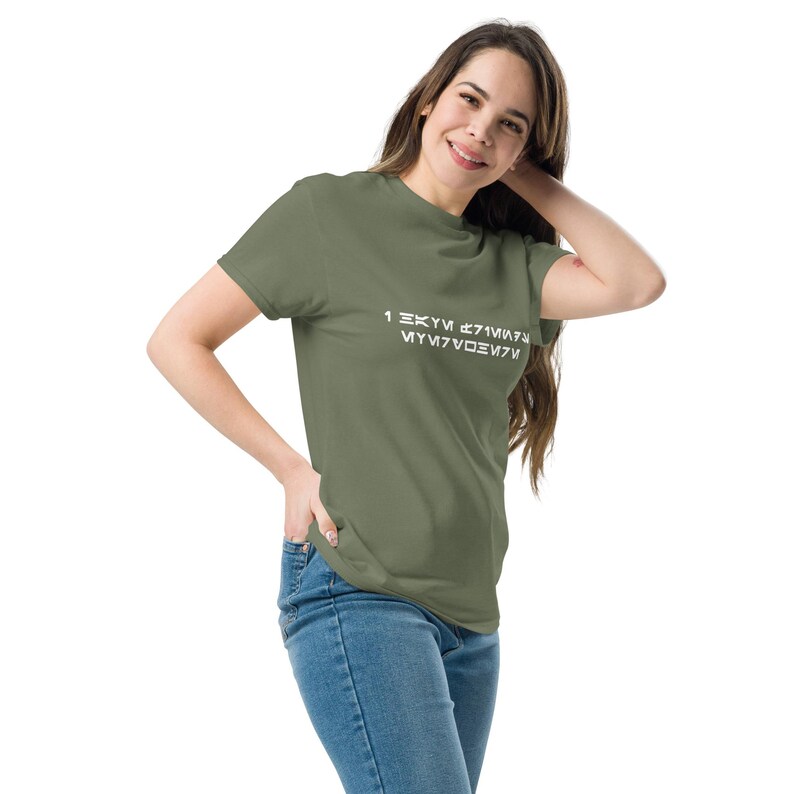 May include: Olive green t-shirt with white text in an alien-like font. The shirt is a crew neck style and is being worn with blue jeans. The model is smiling and has long brown hair.