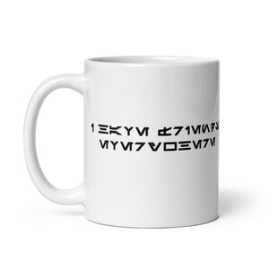May include: White ceramic mug with a black text design. The text appears to be in a stylized, possibly fictional, alphabet. The mug has a standard handle and a glossy finish. A simple, everyday coffee mug.