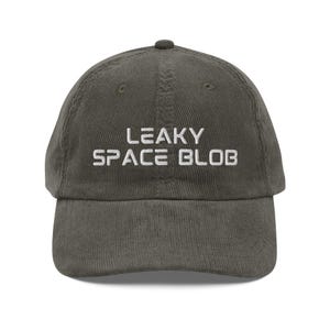 May include: A corduroy baseball cap in a muted olive green color. The cap features the embroidered text "LEAKY SPACE BLOB" in white block letters across the front. The cap has a curved brim and a top button.