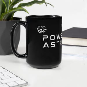 Może przedstawiać: Czarny ceramiczny kubek z białym uchwytem. Kubek zawiera napis "POWER ASTR" pogrubioną czcionką bezszeryfową, wraz z białym symbolem atomu. Kubek stoi na białej powierzchni.