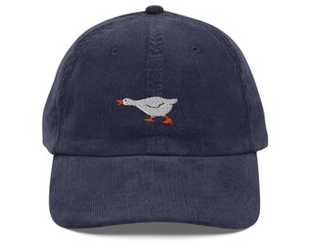 May include: Navy blue corduroy baseball cap featuring an embroidered goose design. The goose is white with orange accents on its beak and feet. The cap has a curved brim and a button on top.