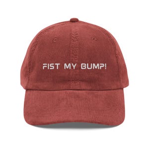 May include: A brick-red corduroy baseball cap with a curved brim. The cap features the embroidered text "FIST MY BUMP!" in white, centered on the front. The hat has a classic six-panel design with a top button.
