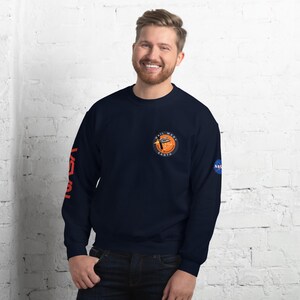 May include: Navy blue sweatshirt with the NASA logo on the right sleeve and a circular patch on the chest that reads "Hail Mary Earth". The left sleeve has the word "NASA" in orange. The sweatshirt is a crew neck style.