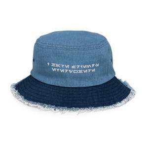 Pode incluir: Um chapéu bucket de ganga em dois tons de azul. A parte superior é azul claro, enquanto a aba é azul mais escuro com uma borda desfiada. Texto branco bordado na frente. Um acessório casual.