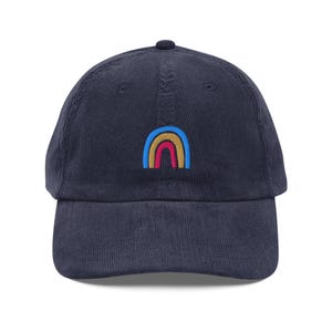 May include: Navy blue corduroy baseball cap with a small embroidered rainbow design in yellow, red, and blue. The cap has a curved brim and a fabric-covered adjustable strap at the back. A casual accessory for everyday wear.