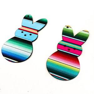 May include: Two wooden Easter bunny ornaments with a colorful striped pattern reminiscent of a Mexican serape. One bunny is blue and green, the other is pink and green.