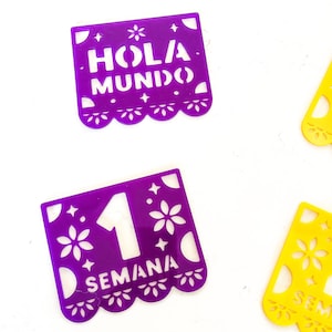 May include: Purple and yellow decorative paper banners with cut-out designs. One purple banner reads "HOLA MUNDO", and another reads "1 SEMANA". The yellow banner has a floral design. These banners are likely for a celebration.