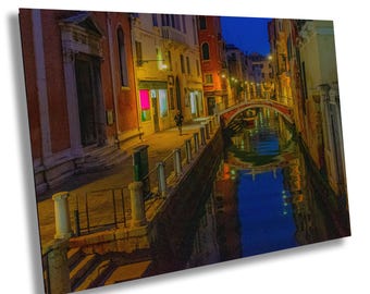 May include: A nighttime view of a Venetian canal with a small bridge and buildings on either side. The water reflects the lights and colors of the buildings and sky. A person walks along the canal.