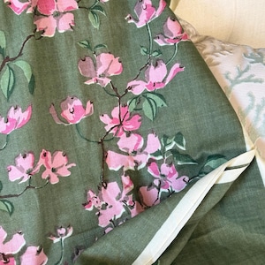 May include: Close-up of a fabric with a floral pattern. The fabric is a muted green color, with pink dogwood blossoms and green leaves. The fabric has a white border.