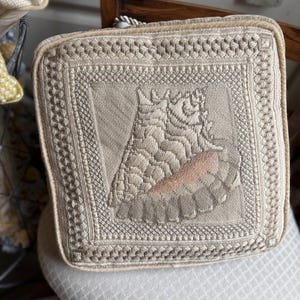 May include: A square, beige embroidered cushion with a detailed floral design in the center. The cushion has a decorative border and a light peach accent in the floral pattern. The cushion is placed on a patterned surface.