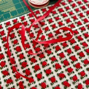 May include: White fabric with a repeating pattern of red and green embroidered flowers. Red ribbon is wrapped around the fabric.