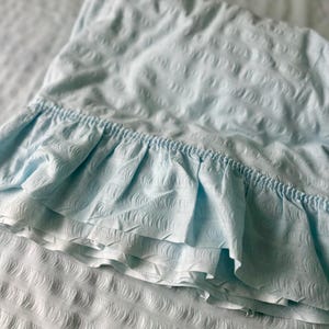 May include: A light blue pillowcase with a ruffled edge, resting on a white bedsheet. The pillowcase and sheet have a textured pattern. The ruffled edge adds a decorative touch to the bedding.