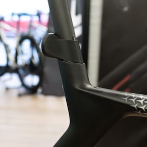 Peut inclure: Gros plan sur un cadre de vélo noir, mettant en évidence la tige de selle et les raccords. Le cadre présente un design moderne avec des motifs en forme d'étoiles. L'arrière-plan est flou, suggérant un magasin ou une exposition.