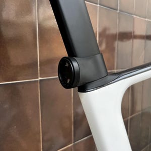 Può includere: Primo piano di un telaio di bicicletta, con reggisella nero e sezione del telaio bianca. Il telaio ha un design moderno e curvo. Il collegamento del reggisella presenta un elemento circolare con piccole viti.
