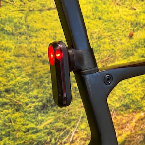 Könnte beinhalten: Ein schwarzes Garmin-Fahrradrücklicht mit rotem Licht ist an einem schwarzen Fahrradrahmen befestigt. Das Licht ist rechteckig mit abgerundeten Kanten. Der Hintergrund ist eine verschwommene Ansicht von grünem Laub.