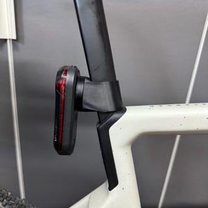 Könnte beinhalten: Ein schwarzes Garmin-Fahrradrücklicht ist an der Sattelstütze eines weißen Fahrradrahmens montiert. Das Licht hat eine rote Linse und ist mit einer schwarzen Halterung befestigt. Der Fahrradrahmen ist weiß mit schwarzen Sprenkeln.
