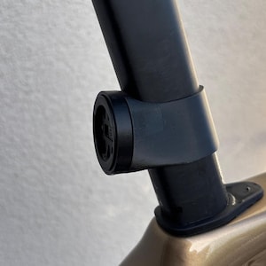 Könnte beinhalten: Nahaufnahme einer schwarzen Fahrradcomputerhalterung, die an einer schwarzen Sattelstütze befestigt ist. Die Halterung ist kreisförmig mit einer Aussparung und einer kleinen schwarzen Halterung. Der Fahrradrahmen ist metallisch goldfarben.