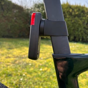 Könnte beinhalten: Ein schwarzes Fahrradrücklicht mit roter Linse ist an einem dunkelgrauen Fahrradrahmen befestigt. Der Fahrradrahmen ist dunkelgrün. Der Hintergrund zeigt verschwommenes grünes Gras und Büsche.
