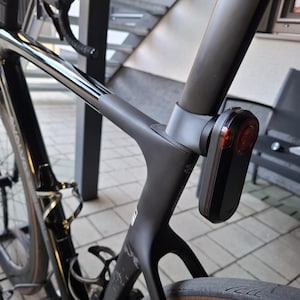 Op de afbeelding: Een zwarte racefiets met een rood achterlicht bevestigd aan de zadelpen. De fiets heeft een gestroomlijnd frame en een bidonhouder. De banden hebben een bruine zijwand.