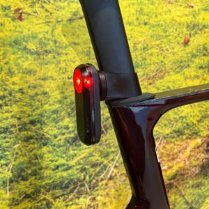 Peut inclure: Un feu arrière de vélo Garmin noir avec deux lumières rouges est monté sur un cadre de vélo rouge foncé. La lumière est fixée à la tige de selle. L'arrière-plan est une vue floue de feuillage vert.