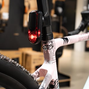 Può includere: Primo piano di una bicicletta con telaio a fantasia rosa e nero. Una luce rossa per bici Garmin è attaccata al reggisella. Lo pneumatico della bicicletta è nero.