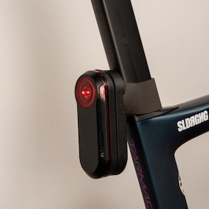 Könnte beinhalten: Schwarzes Garmin-Fahrradrücklicht mit roter Linse, befestigt an einem dunkelblauen Fahrradrahmen. Das Licht ist an der Sattelstütze des Fahrrads montiert. Der Fahrradrahmen trägt das Wort "Tarmac".