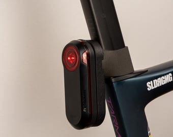 May include: Black Garmin bike light with a red lens attached to a dark blue bicycle frame. The light is mounted on the seat post of the bike. The bike frame has the word "Tarmac" printed on it.