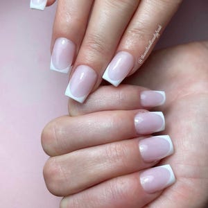 Op de afbeelding: Close-up van handen met korte, vierkante nagels die zijn gelakt met een lichtroze lak en een witte french tip.
